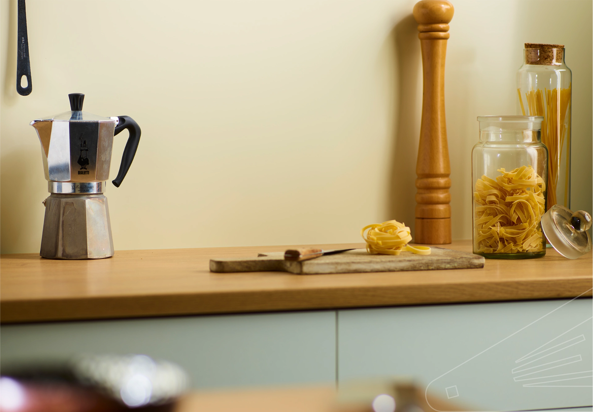 Ivory Satin Cladding installed above a wooden kitchen worktop. The surface is holding a chopping board, glass storage jars and a coffee percolator.