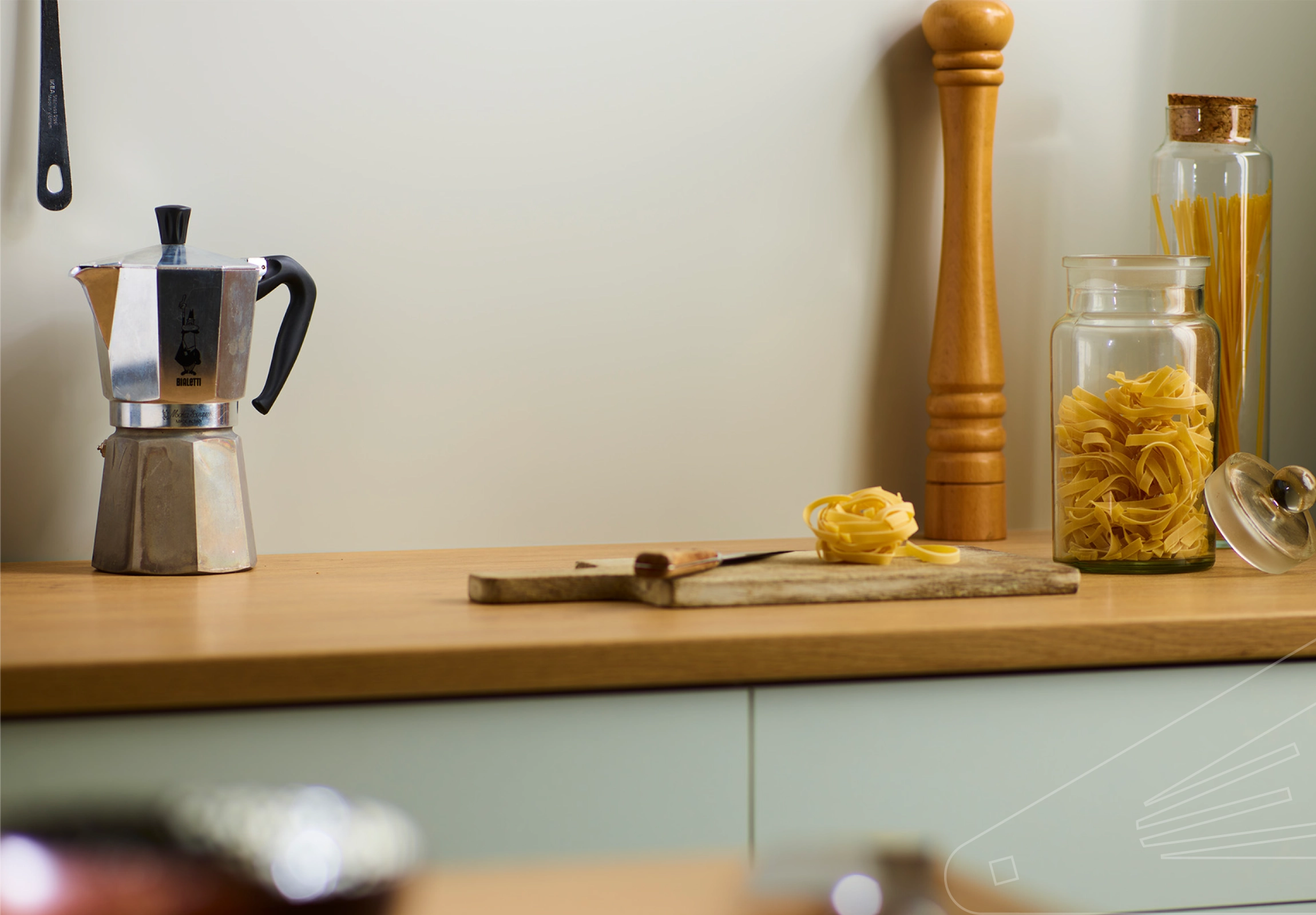 Pebble Satin Cladding installed above a wooden kitchen worktop. The surface is holding a chopping board, glass storage jars and a coffee percolator.