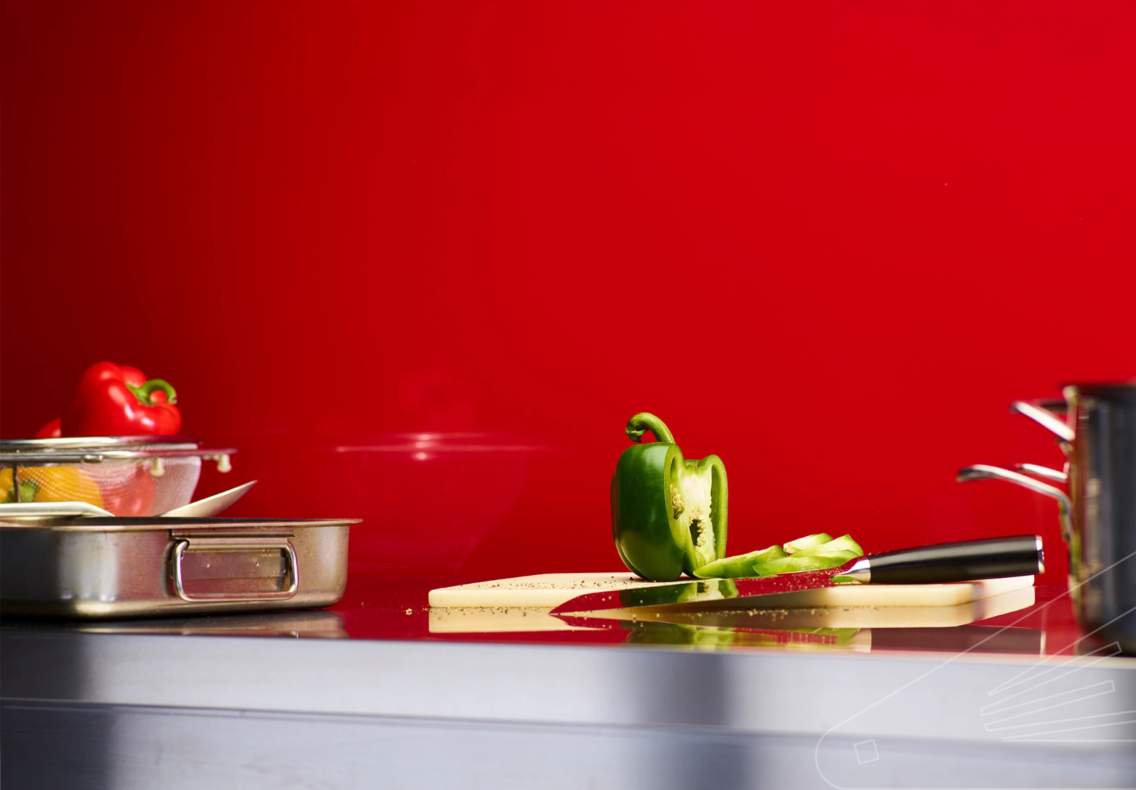 Ruby Gloss Cladding installed above a stainless steel kitchen worktop. The surface is holding a chopping board, pans, utensils, and various colourful peppers.