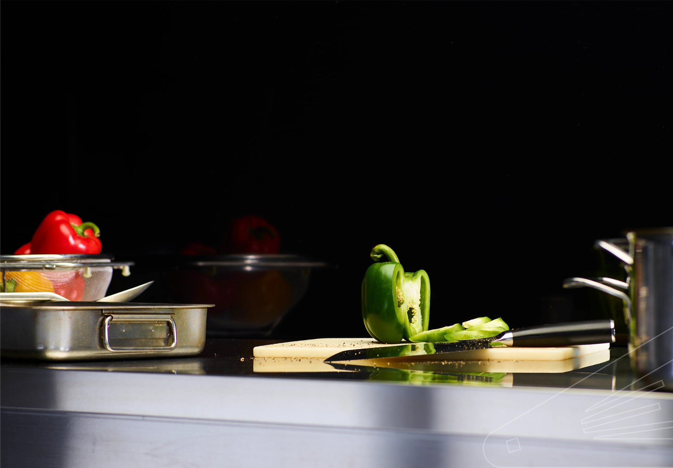 Slate Gloss Cladding installed above a stainless steel kitchen worktop. The surface is holding a chopping board, pans, utensils and various colourful peppers.